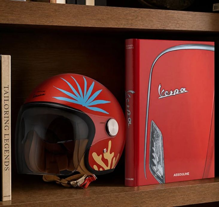 A red Vespa helmet with blue and gold motifs next to the red Assouline Vespa book on a wooden shelf