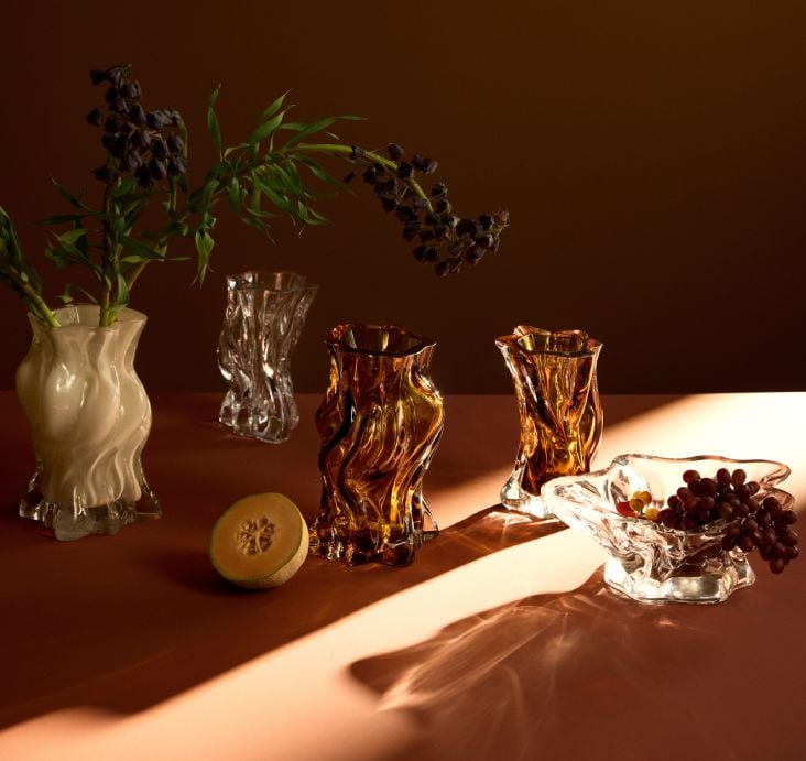 Four AYTM 'Vena' mouthblown glass vases in amber and crystal tones with flowers on a warm terracotta surface