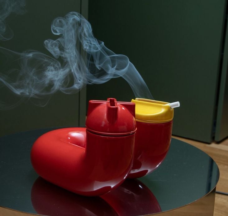 Two red Artetica 'U' ceramic ashtray add-ons with smoke rising from one, on a dark round table