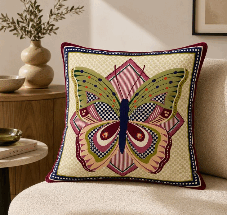 A Vesta cushion with a vivid embroidered butterfly motif on a cream armchair next to a side table